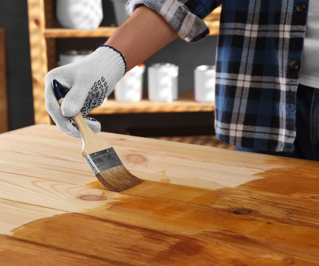 Holzoberfläche wird mit einem Pinsel und Holzschutzlasur gestrichen, Hand mit Arbeitshandschuh trägt gleichmäßig Holzöl auf.