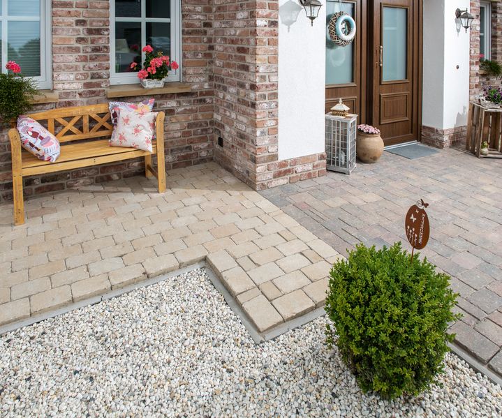 Hauseingang mit gemischtem Pflasterbelag in Sandstein- und Grautönen, Sitzbank, Kiesfläche und bepflanztem Vorgarten.