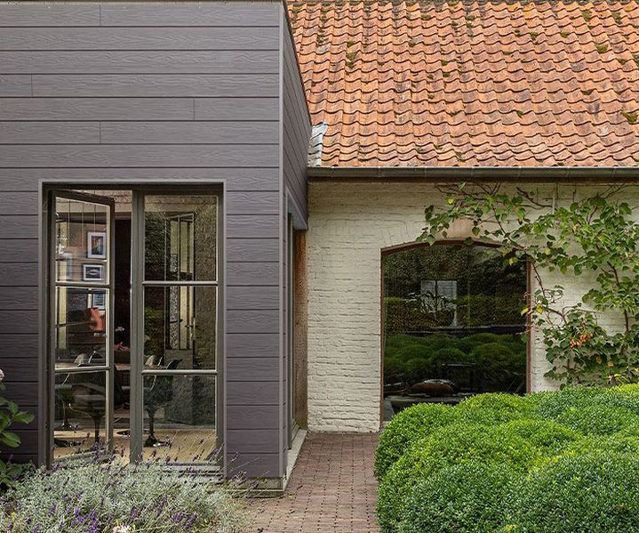 Gebäudefassade mit moderner, grauer Fassadenverkleidung im Holzlook neben einem bestehenden Backsteingebäude mit rotem Ziegeldach. Der Übergang zwischen Alt- und Neubau ist klar und hochwertig gestaltet.