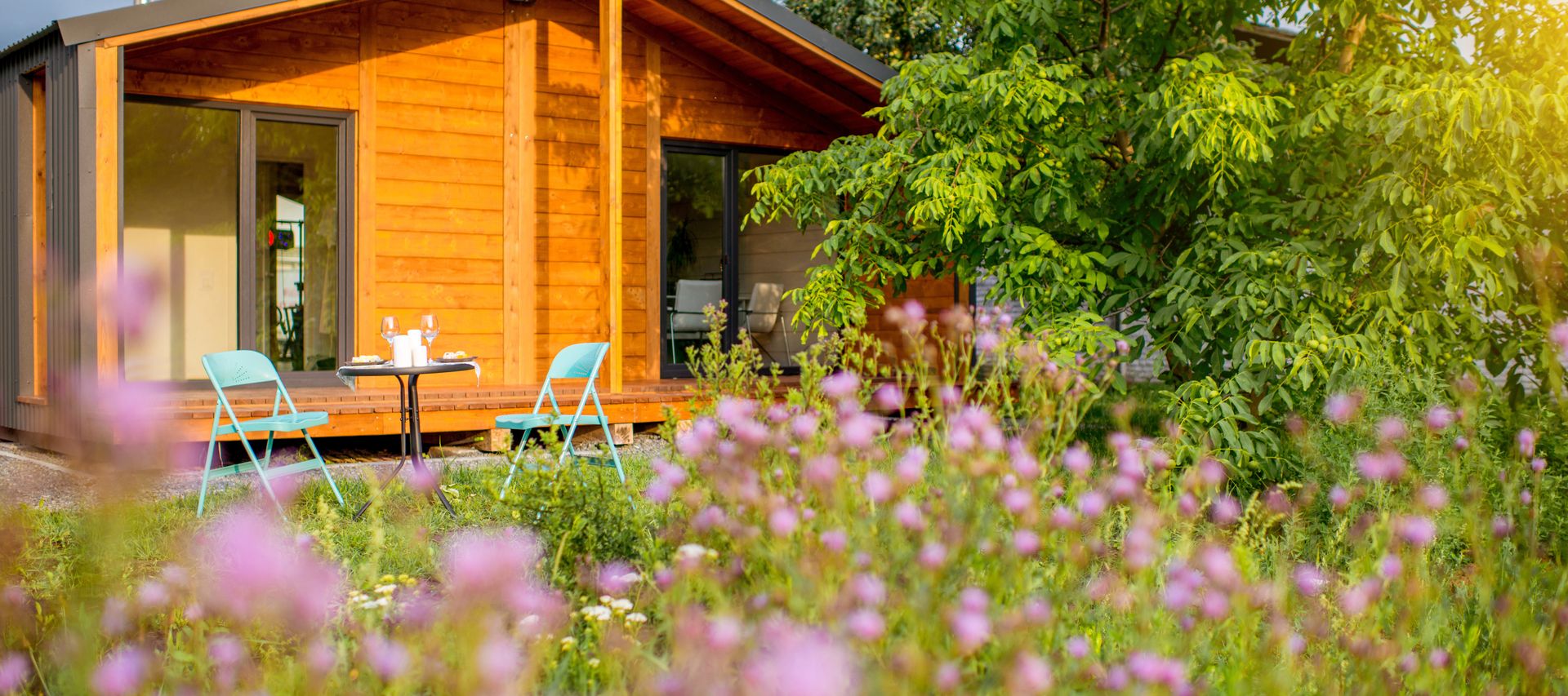 Modernes Gartenhaus aus Holz mit Terrasse, zwei Stühlen und Tisch, umgeben von blühender Wiese und Bäumen im warmen Sonnenlicht