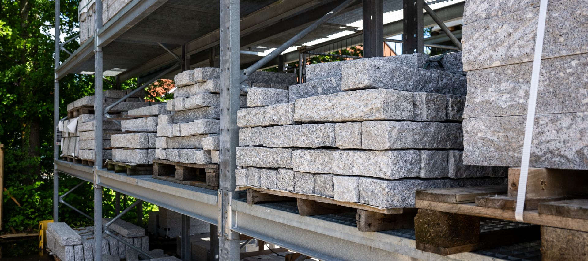 Gestapelte Natursteinplatten und Mauersteine lagern sortiert in einem Außenregal im Baustoffhandel.