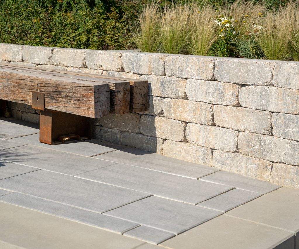 Sitzbank aus massivem Holz vor einer Trockenmauer aus hellen Natursteinblöcken, kombiniert mit großformatigen Terrassenplatten und Gräserbepflanzung.