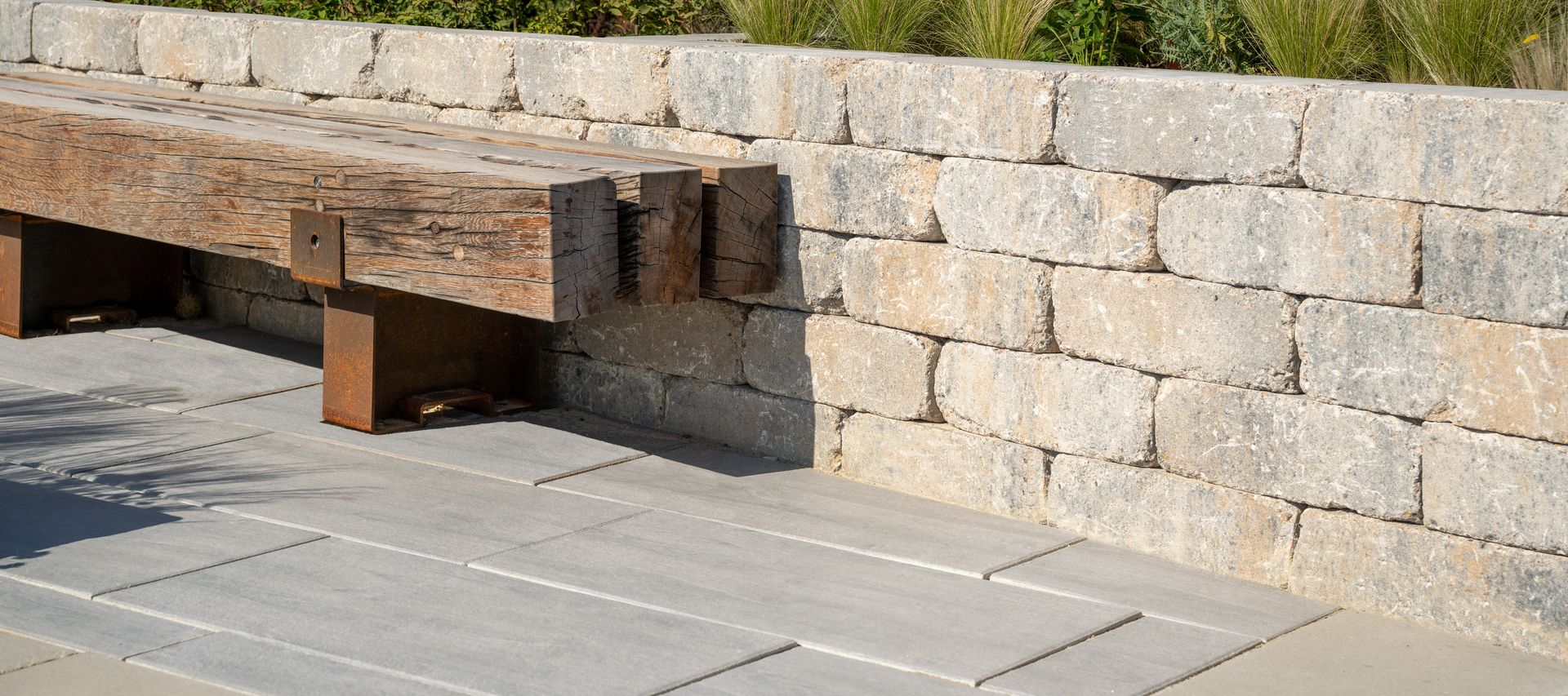 Sitzbank aus massivem Holz vor einer Trockenmauer aus hellen Natursteinblöcken, kombiniert mit großformatigen Terrassenplatten und Gräserbepflanzung.