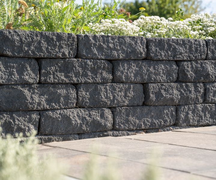 Dunkelgraue Trockenmauer aus rustikal strukturierten Mauersteinen mit bepflanzter Oberkante und angrenzender Terrasse aus Betonplatten.
