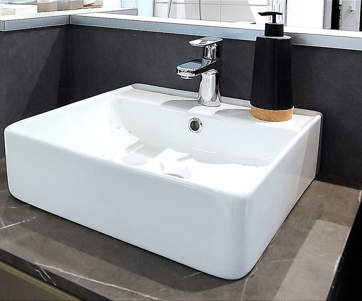 Modernes weißes quadratisches Waschbecken mit silbernem Wasserhahn auf dunkler Arbeitsplatte, begleitet von einem schwarzen Seifenspender.