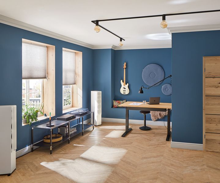 Modernes Zimmer mit blauen Wänden, Holzboden, Schreibtisch und Gitarre ausgestellt. Sonnenlicht strömt durch Fenster mit Jalousien und schafft so eine gemütliche Atmosphäre.