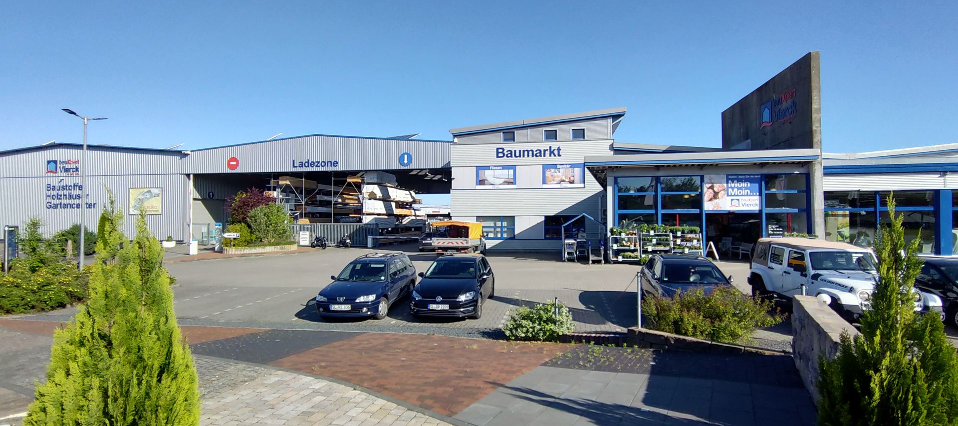Ein Baumarkt mit Parkplatz, mehreren geparkten Autos und klarem blauen Himmel. Im Vordergrund stehen grüne Sträucher und gepflasterte Gehwege.