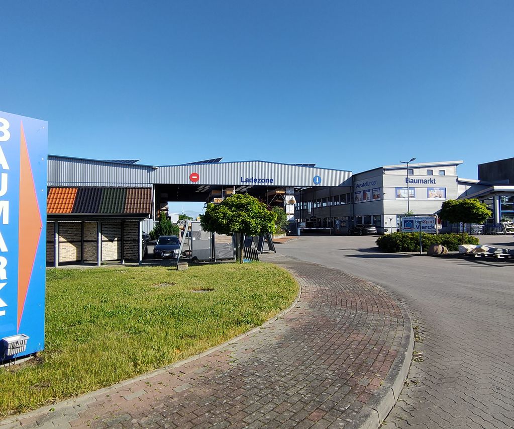 Außenansicht eines Baumarkts mit „Baumarkt“-Schild, Ladezone und umliegenden gepflasterten und grasbewachsenen Flächen unter klarem blauen Himmel.