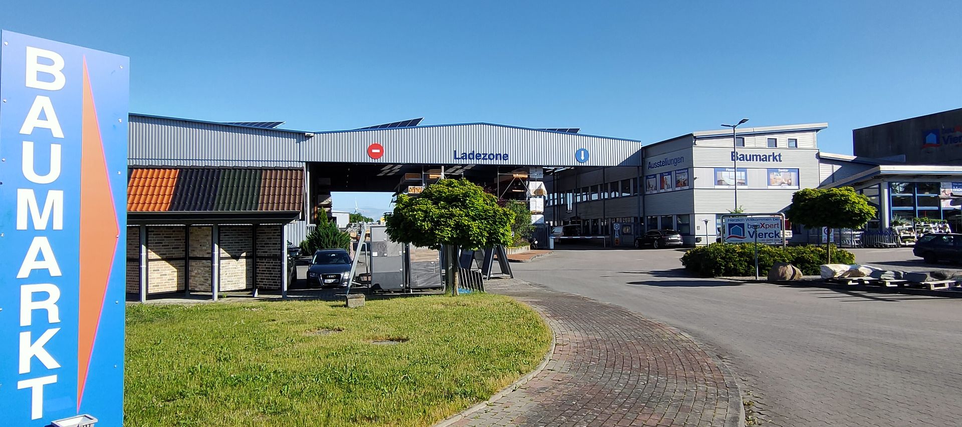 Außenansicht eines Baumarkts mit „Baumarkt“-Schild, Ladezone und umliegenden gepflasterten und grasbewachsenen Flächen unter klarem blauen Himmel.
