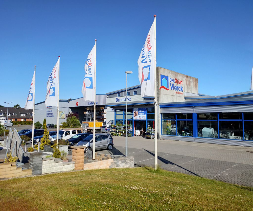 Außenansicht eines modernen Baumarkts mit blauen Akzenten, umgeben von Flaggen mit Firmenloge bauXpert Vierck und geparkten Autos, unter klarem blauen Himmel.