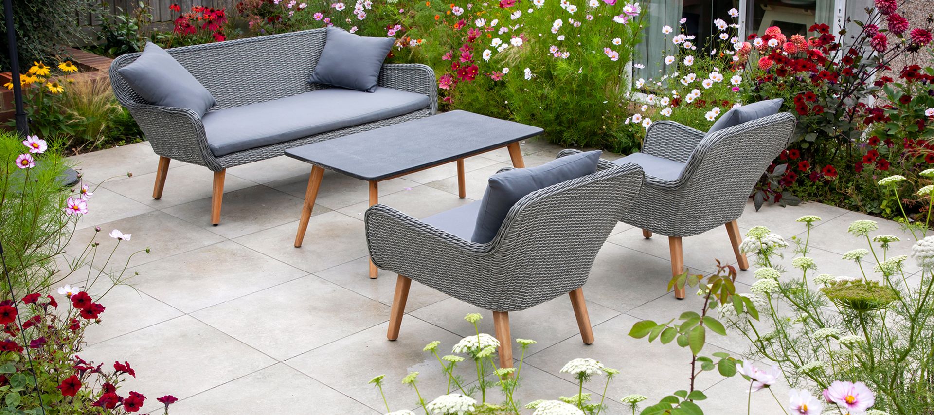 Terrasse mit hellen Keramikterrassenplatten, grauen Gartenmöbeln und blühender Bepflanzung im umliegenden Garten.