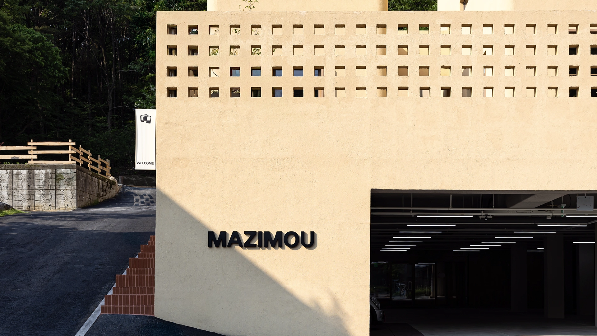 Modern MAZIMOU building with a textured beige facade featuring square cutout windows and large black 'MAZIMOU' lettering at the entrance.