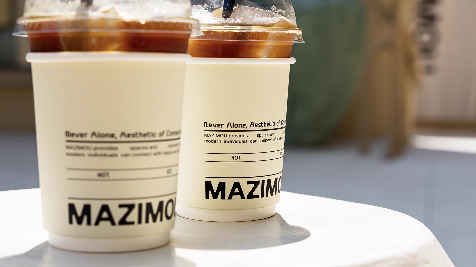 Two iced coffee cups from MAZIMOU on a white table, featuring 'Never Alone, Aesthetic of Coexistence' and 'MAZIMOU' branding.