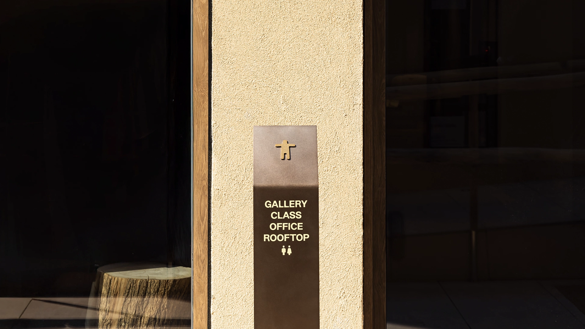 Close-up of a dark metal signpost at MAZIMOU indicating 'GALLERY CLASS OFFICE ROOFTOP' with directional pictograms, next to a glass door.