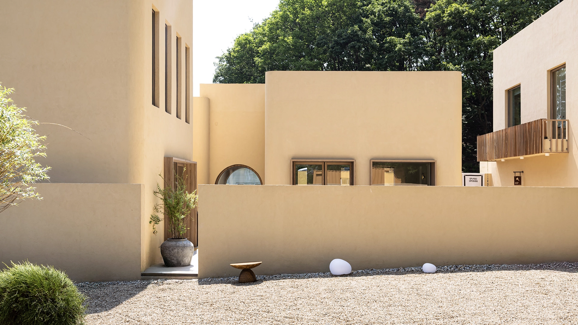 Exterior view of MAZIMOU, a contemporary building complex with light beige walls and various window shapes, surrounded by gravel and trees.