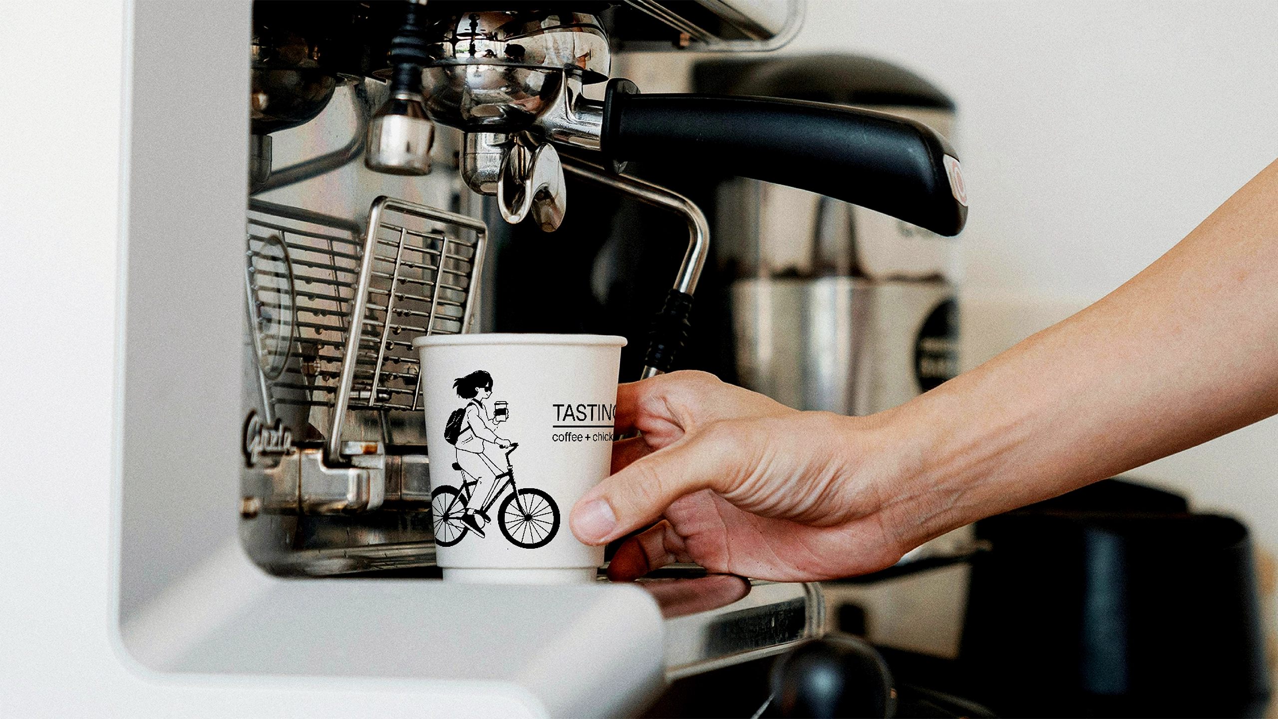 Hand holding a branded coffee cup with a bicycle illustration, next to an espresso machine