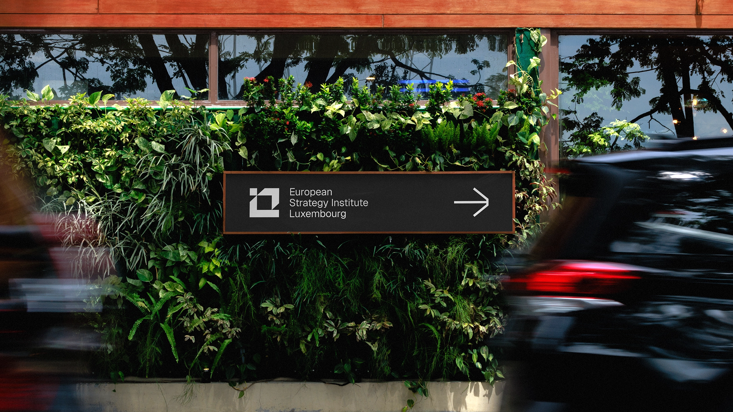 European Strategy Institute Luxembourg wayfinding sign mounted on a lush green living plant wall, featuring white logo and directional arrow on dark charcoal background with copper frame