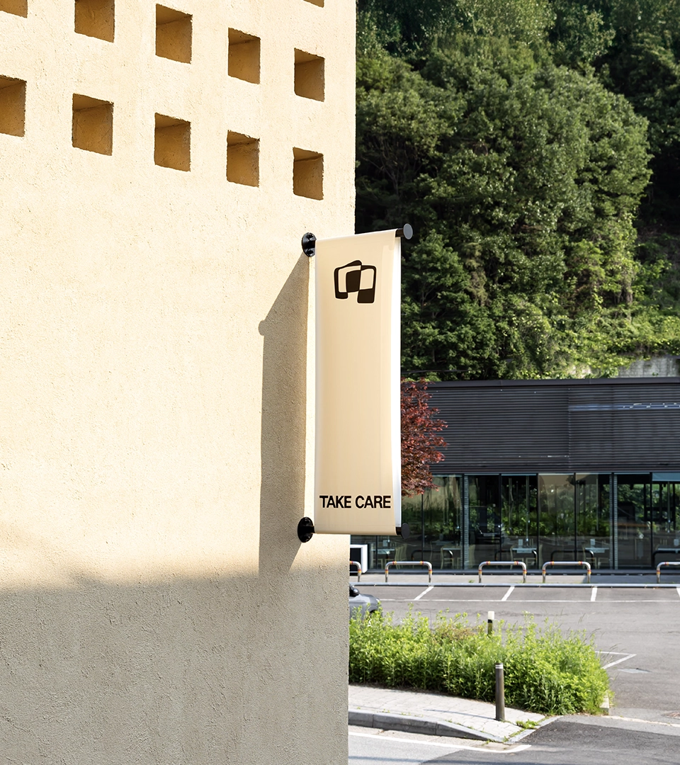 MAZIMOU building exterior with a tall vertical banner displaying a logo and the text 'TAKE CARE', overlooking a parking lot and greenery.