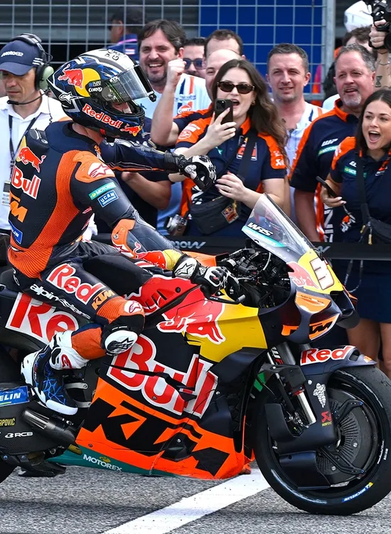 Pedro Acosta on a Red Bull KTM bike greets cheering fans and photographers at a racing event, with people capturing the moment energetically.