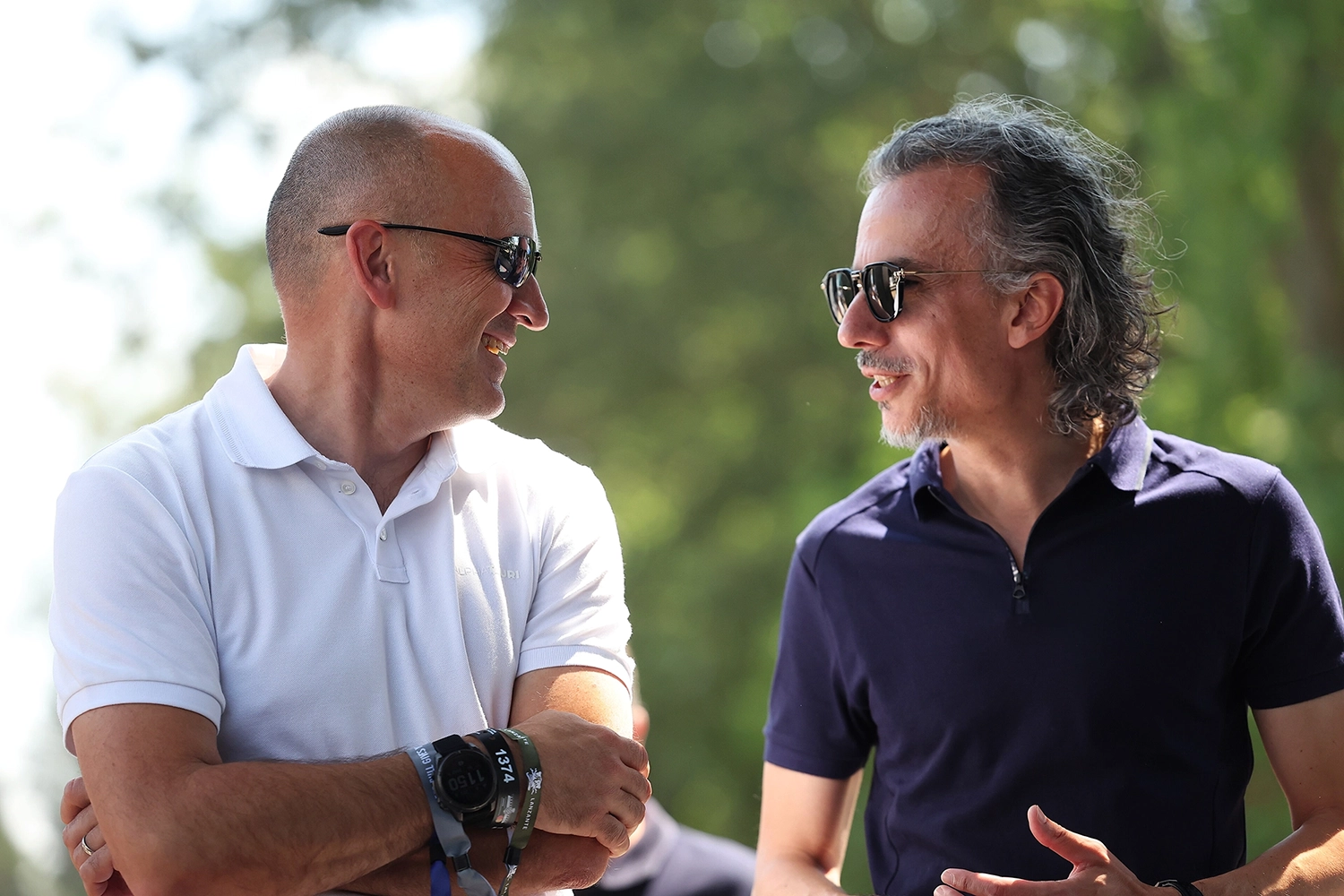 Two men wearing sunglasses engaged in conversation outdoors, one in a white shirt and the other in a navy shirt, with trees in the background.