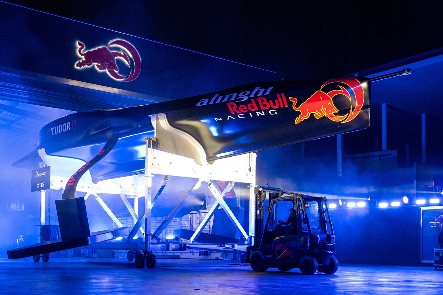 A racing sailboat being hoisted indoors, featuring Alinghi Red Bull Racing logos, illuminated by blue lights.