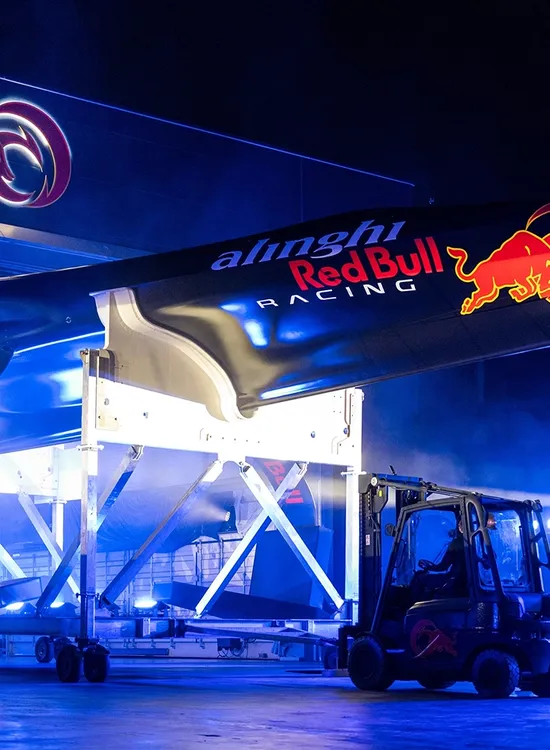 A racing sailboat being hoisted indoors, featuring Alinghi Red Bull Racing logos, illuminated by blue lights.