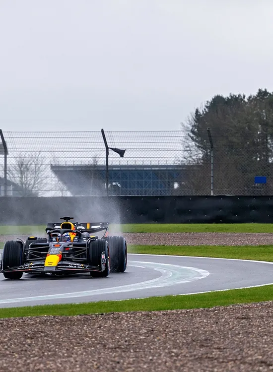 A Formula 1 car racing on a wet track, creating spray behind it as it rounds a curve, with a trackside barrier and leafless trees in the background.