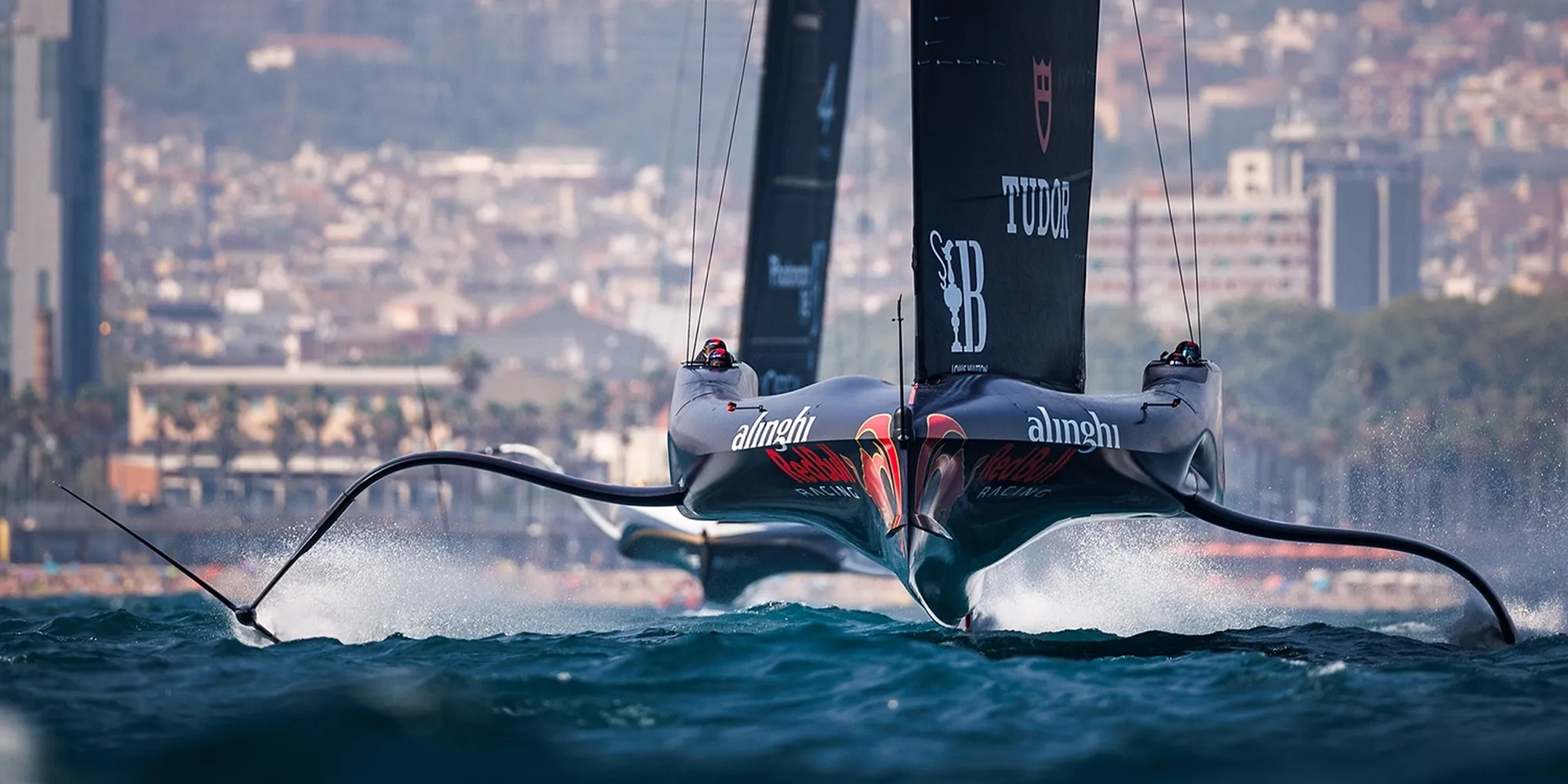 Sleek sailboats with foils skimming over choppy water near a city shoreline in the background.