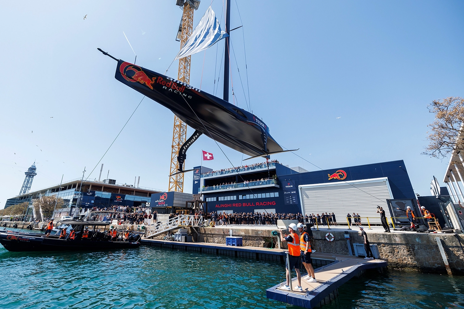 A sailboat with Red Bull Racing branding is lifted by a crane in a harbor, surrounded by people on docks and a boat on the water.
