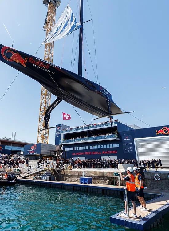 A sailboat with Red Bull Racing branding is lifted by a crane in a harbor, surrounded by people on docks and a boat on the water.