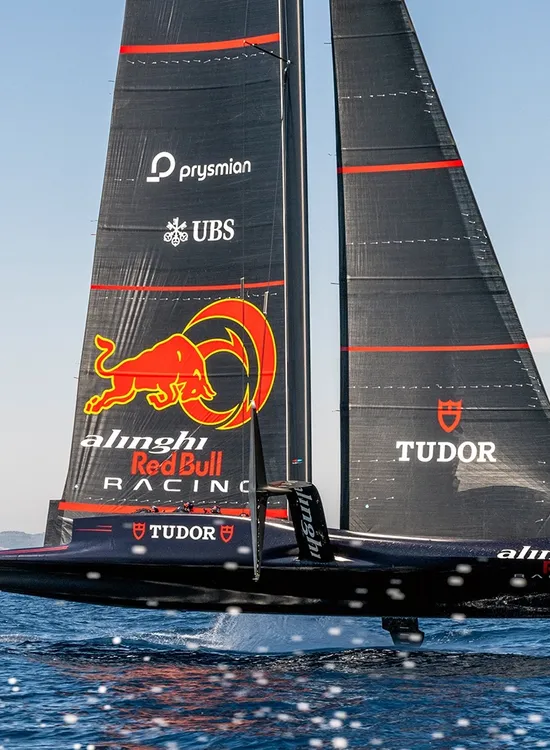 A sleek Alinghi Red Bull Racing sailboat with dark sails glides over the water, showcasing logos against a cityscape backdrop.