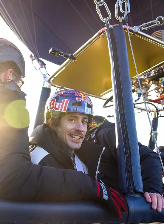 A person wearing a helmet and gloves smiles inside a hot air balloon basket, with sunlight streaming in from the left. Others with helmets are visible.