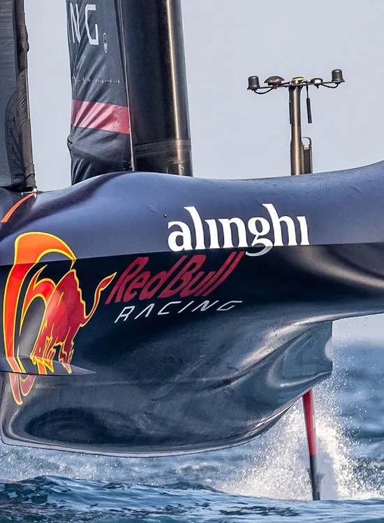 A high-speed racing sailboat with "Alinghi" and "Red Bull Racing" logos skims the water's surface, with crew members visible wearing helmets.