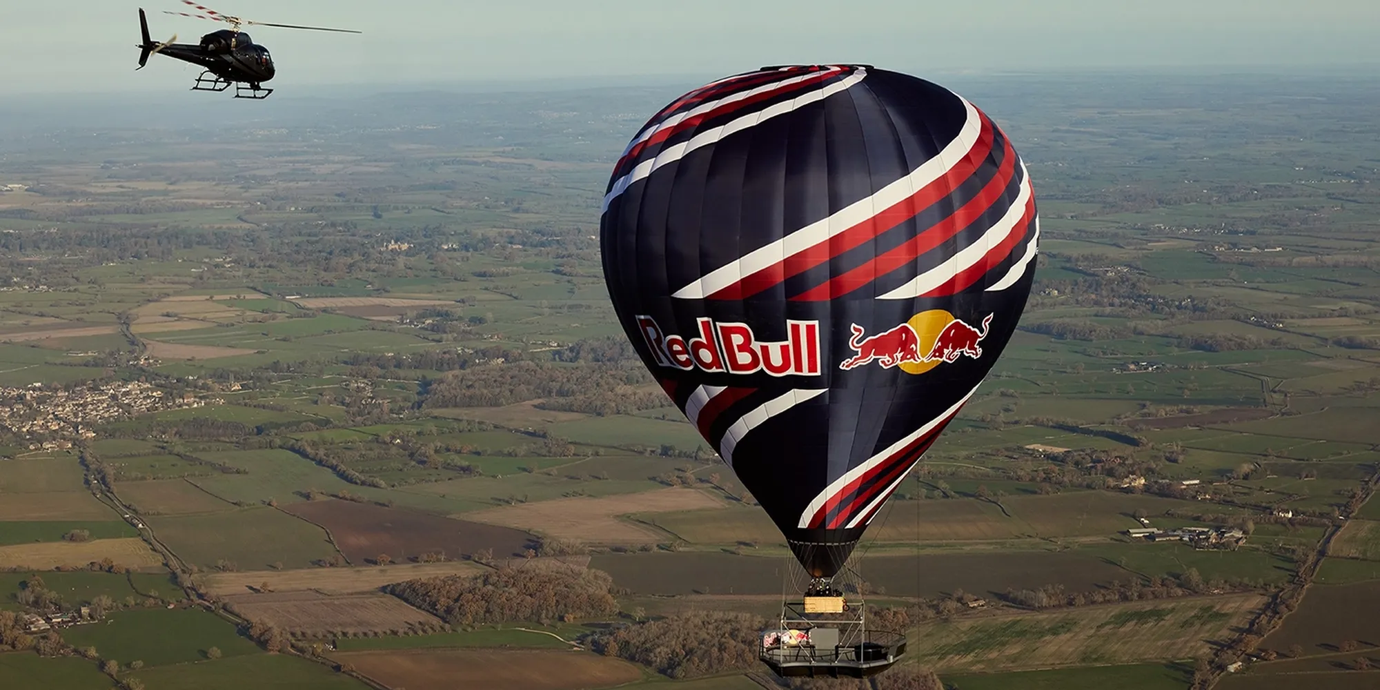 A helicopter flies near a hot air balloon with Red Bull branding, soaring above a sprawling green landscape with fields and trees.
