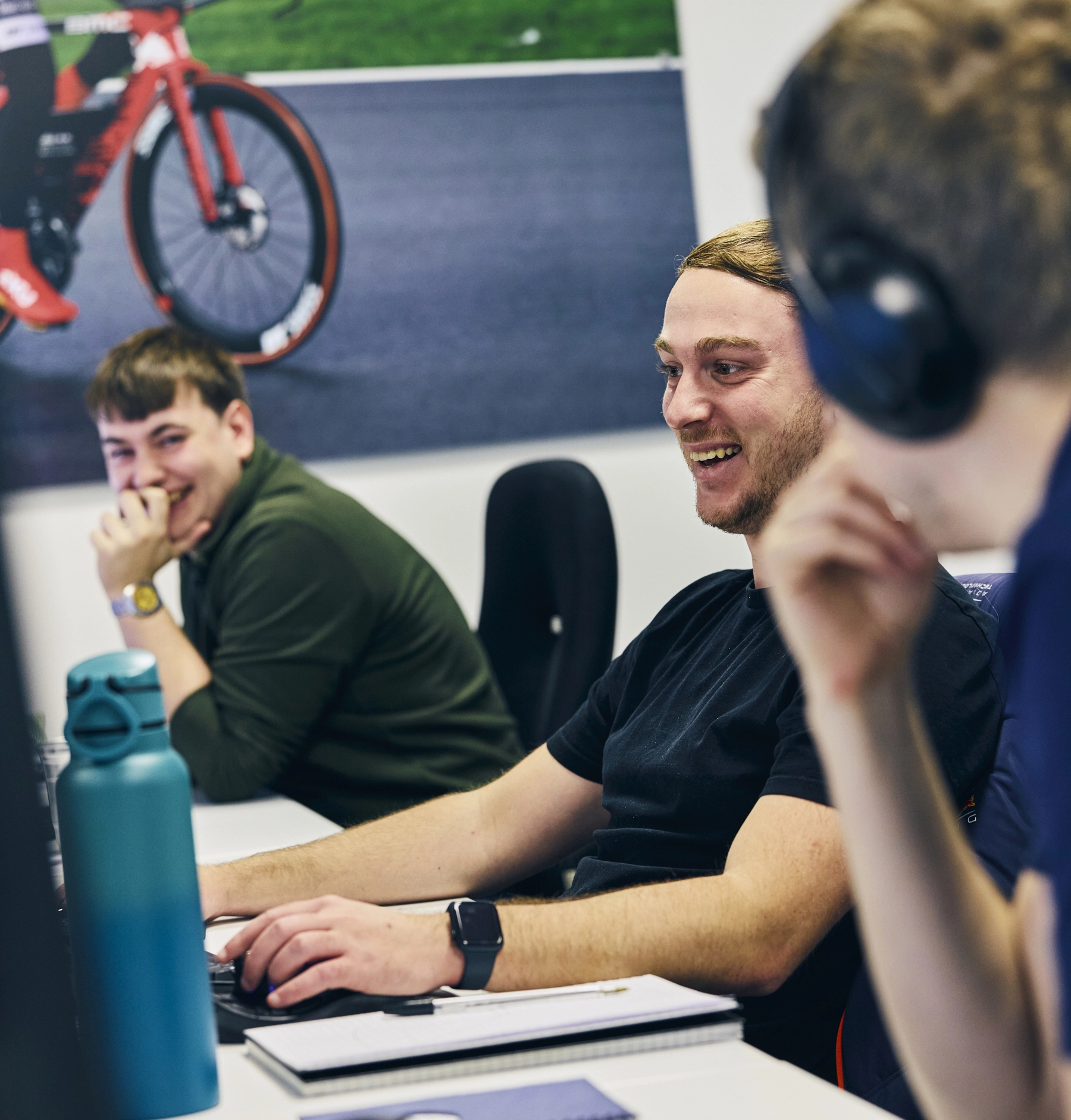 Three people sitting at desks working with laptops, smiling and talking. A bike poster is visible in the background.