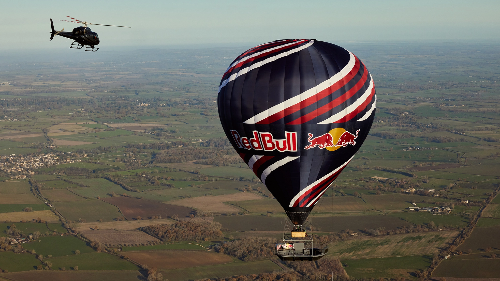 A large hot air balloon with "Red Bull" logo flies over a rural landscape, accompanied by a helicopter.