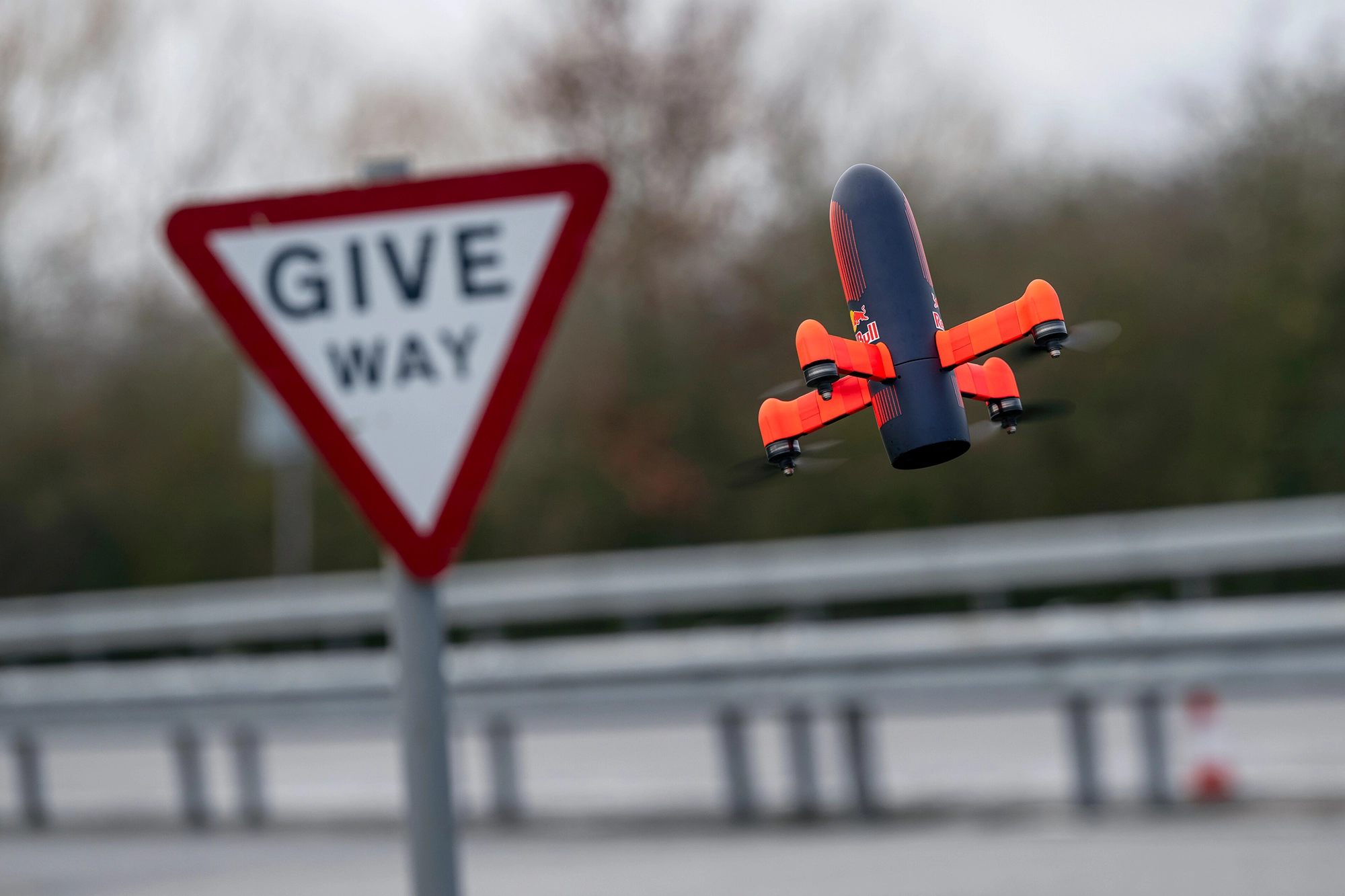 A small orange and black drone flies near a "Give Way" road sign with a blurred background of trees and a metal guardrail.
