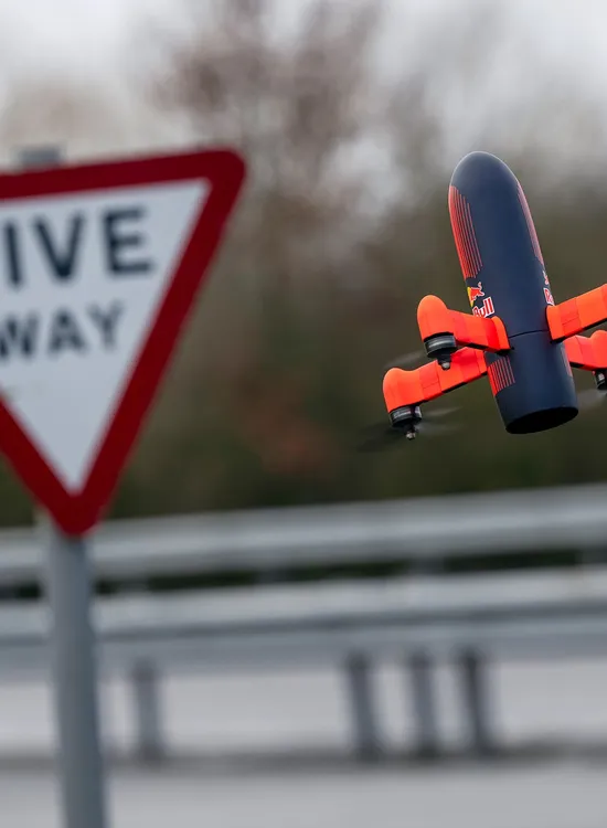 A small orange and black drone flies near a "Give Way" road sign with a blurred background of trees and a metal guardrail.