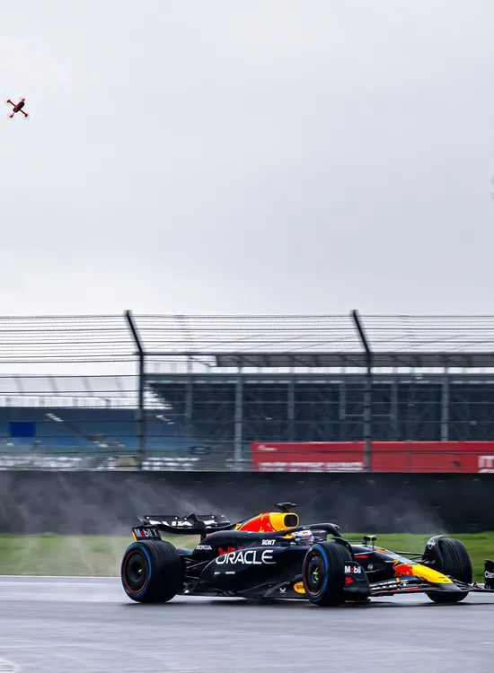 A Formula 1 car speeds along a wet racetrack, creating spray, with overcast skies and a bare tree visible in the background.