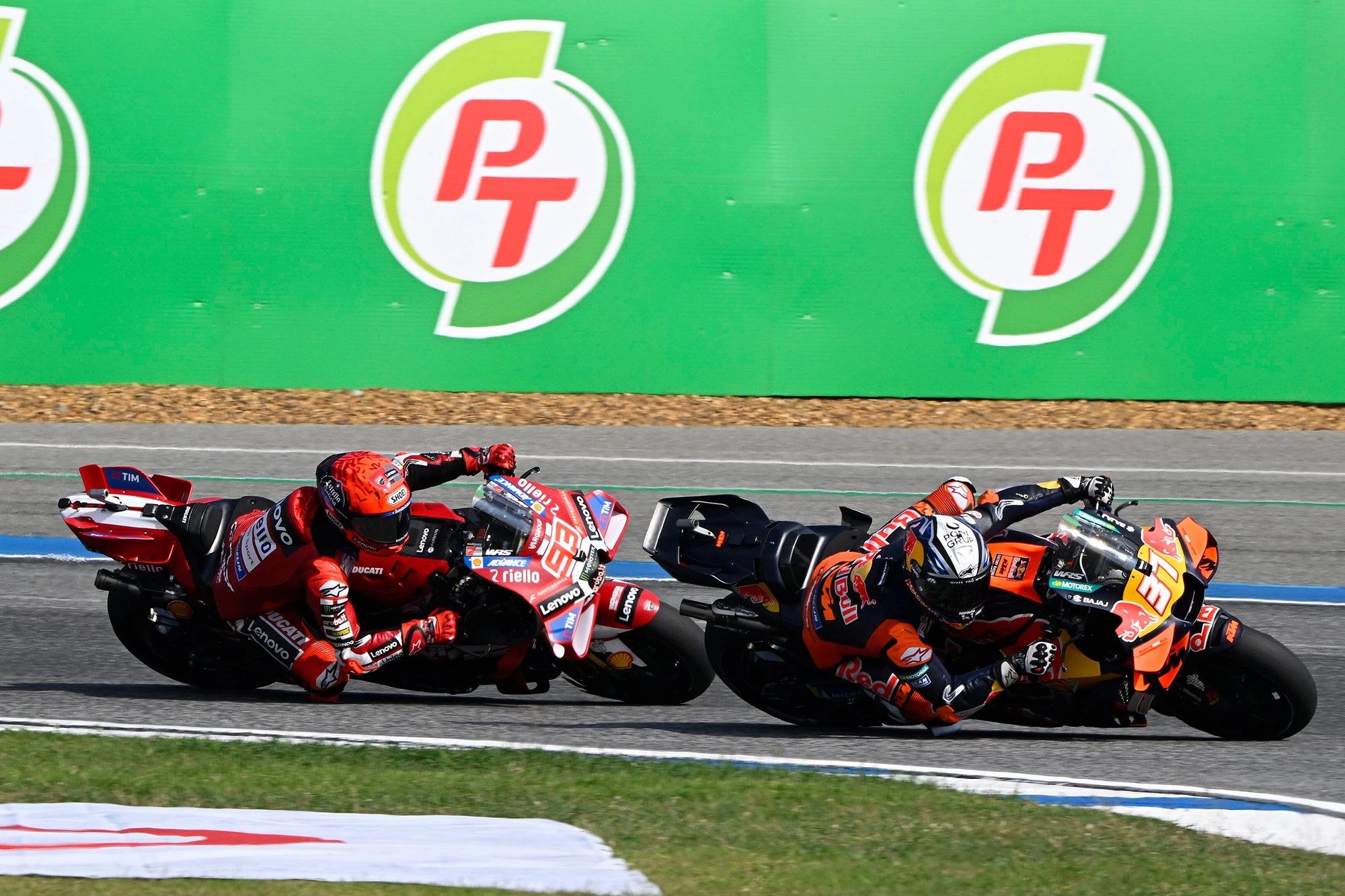 Two motorcycle racers in red and orange gear lean into a sharp turn on a track, with green advertising boards in the background.