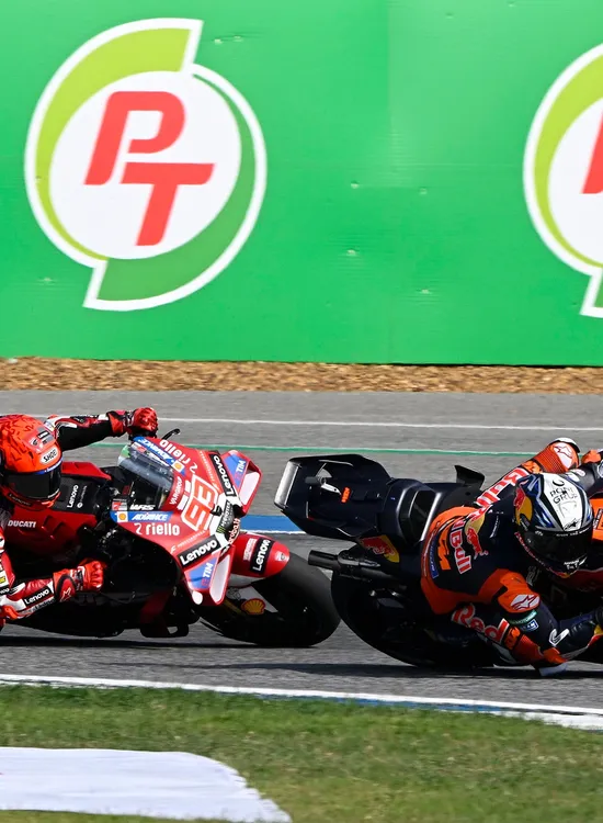Two motorcycle racers in red and orange gear lean into a sharp turn on a track, with green advertising boards in the background.