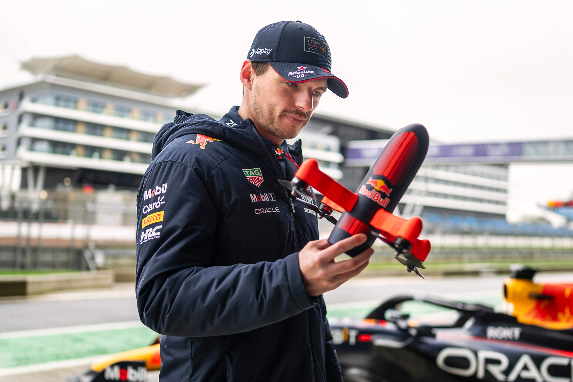 Max Verstappen holds a drone, standing by a circuit with modern buildings in the background.