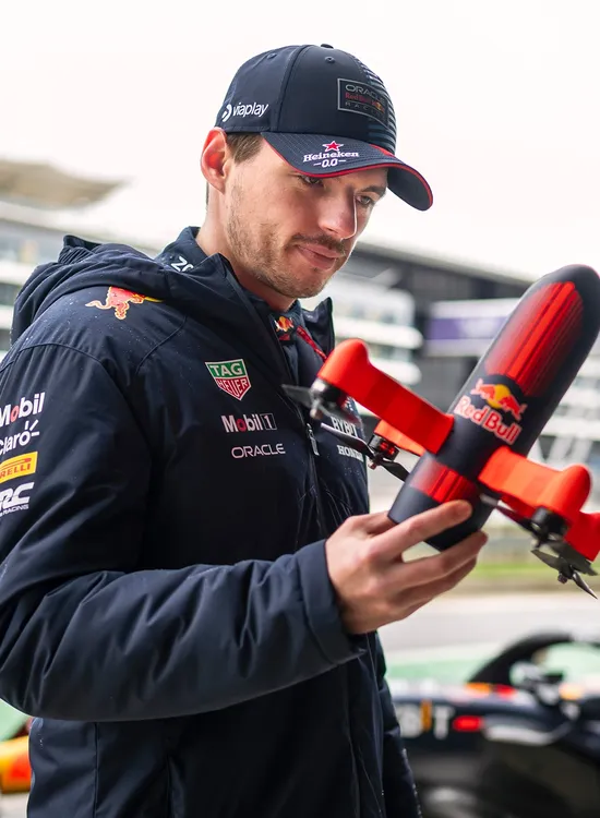 Max Verstappen holds a drone, standing by a circuit with modern buildings in the background.
