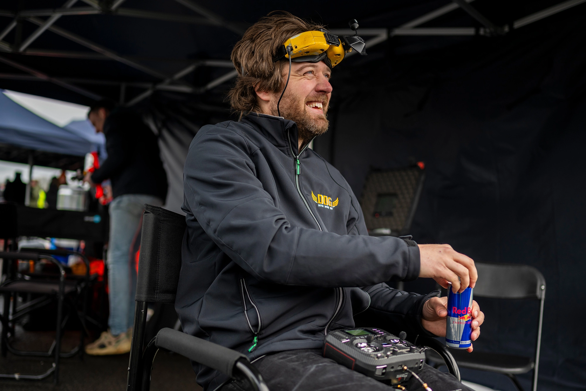 Shaggy FPV Ralph Hogenbrik smiles while holding a can of Red Bull. They are wearing a jacket and FPV goggles under a tent with equipment around.
