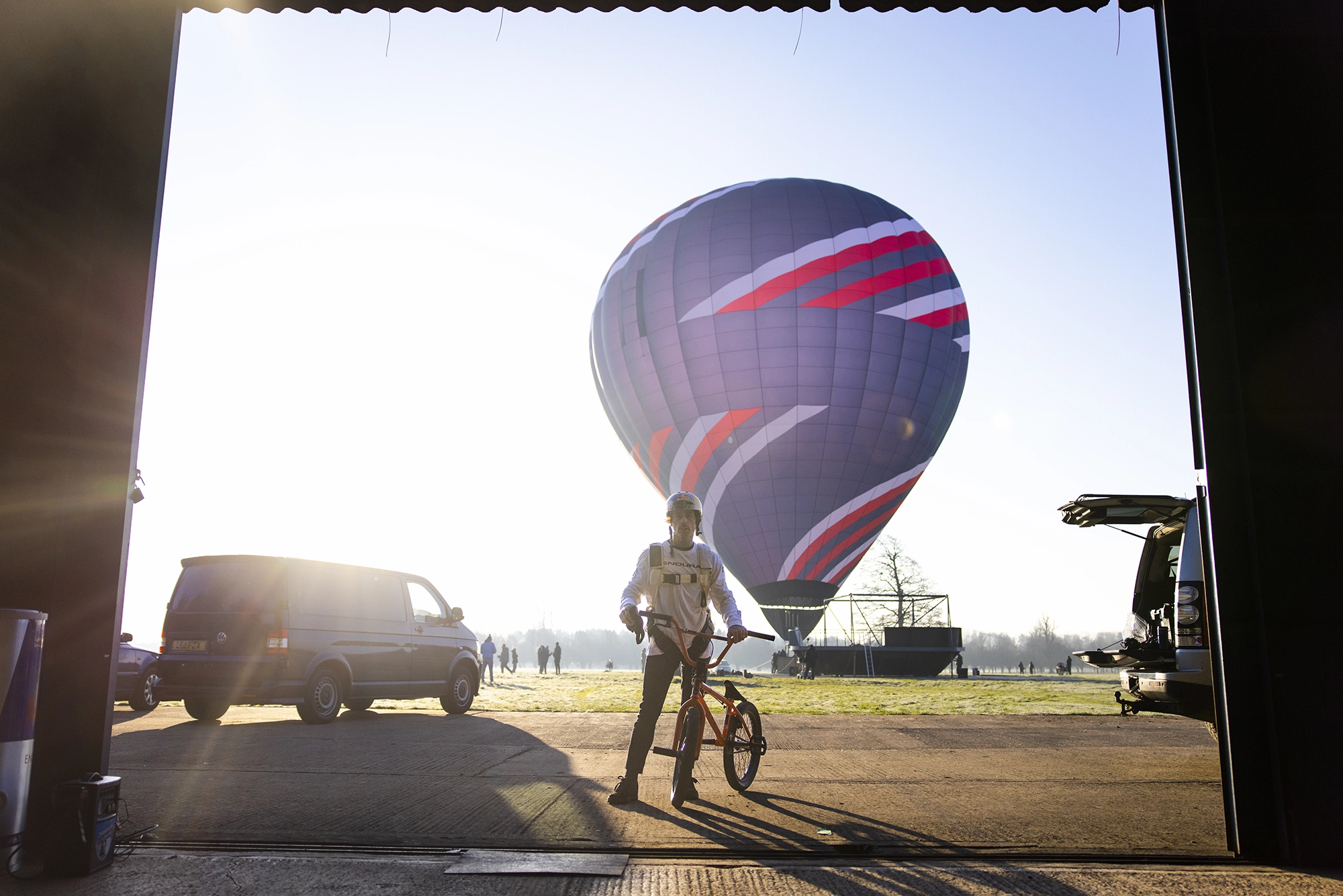 Kris Kyle with a BMX bike stands in a hangar, silhouetted against a rising sun. Outside, a hot air balloon and vehicles are visible.