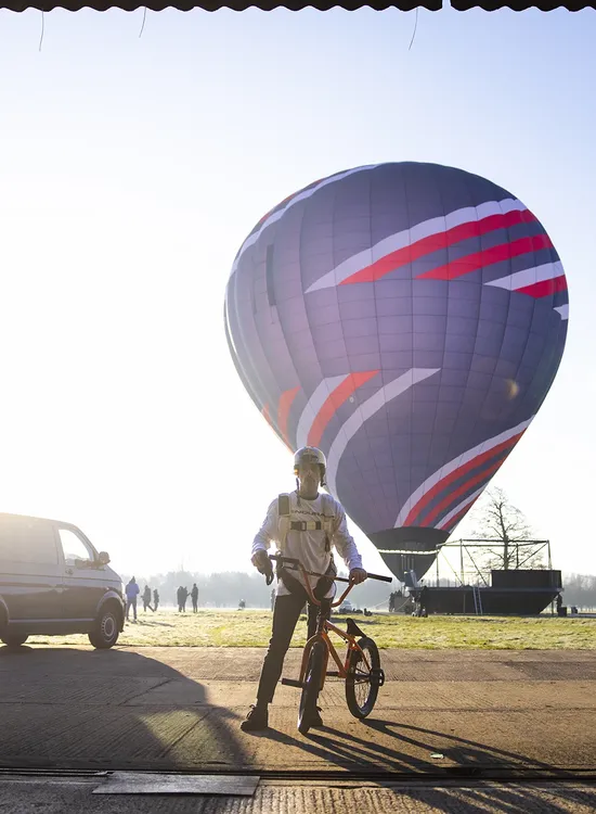 Kris Kyle with a BMX bike stands in a hangar, silhouetted against a rising sun. Outside, a hot air balloon and vehicles are visible.