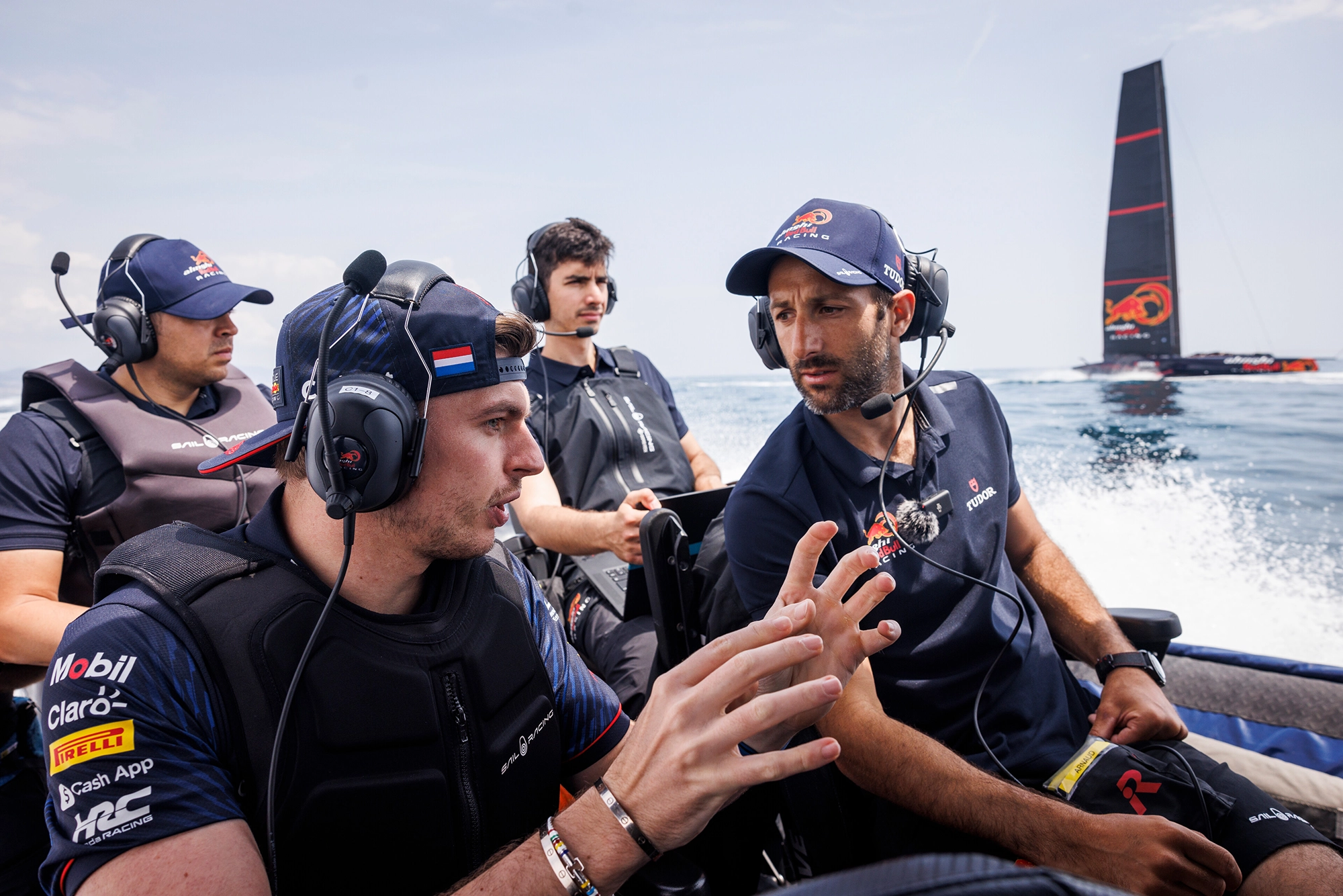 Four men in Red Bull racing gear converse on a speedboat during a race, with a large sailboat visible in the background.