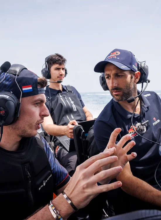 Four men in Red Bull racing gear converse on a speedboat during a race, with a large sailboat visible in the background.