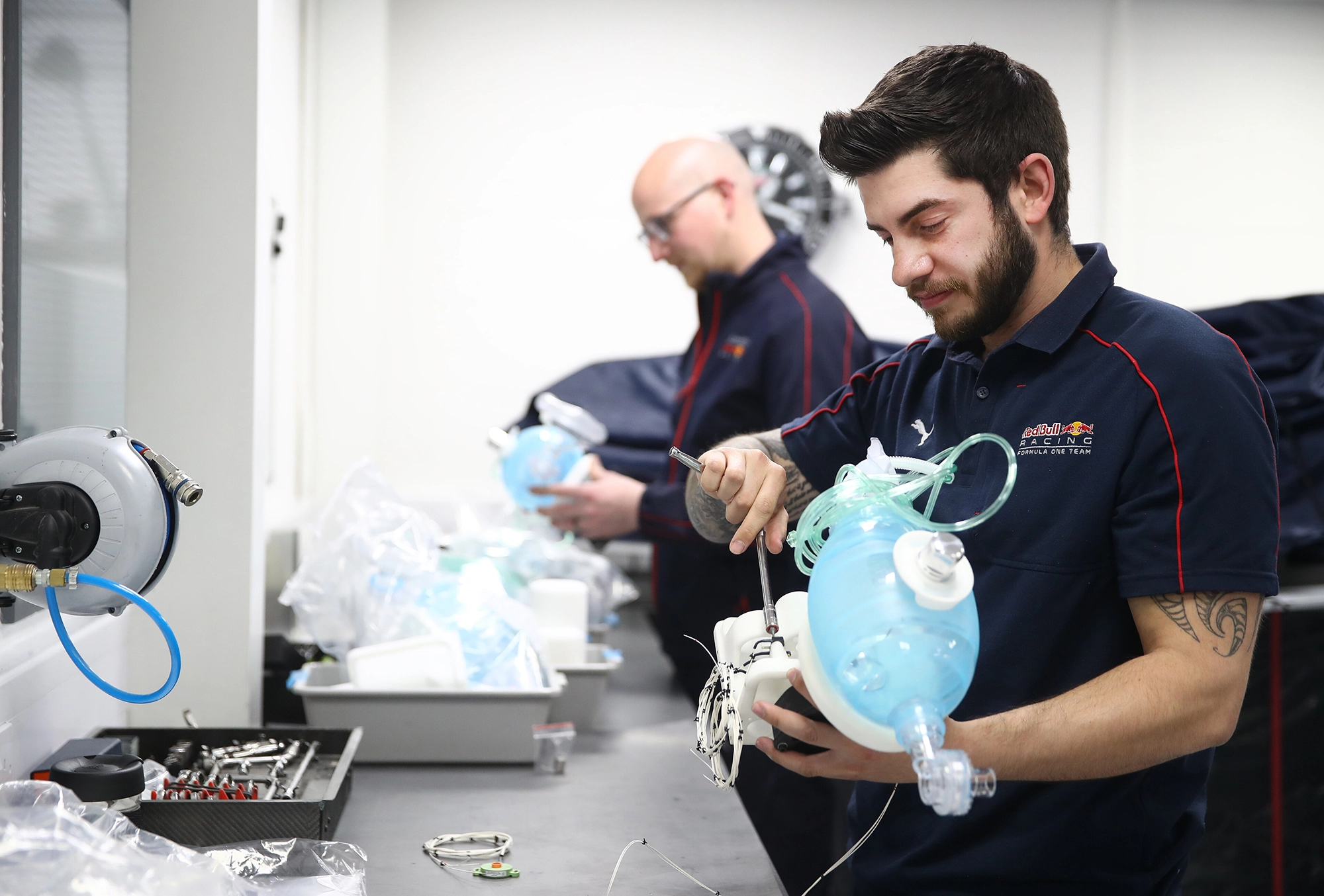 Two men in uniform assemble medical ventilators at a workshop, working with blue ventilator bags and tools on a table.