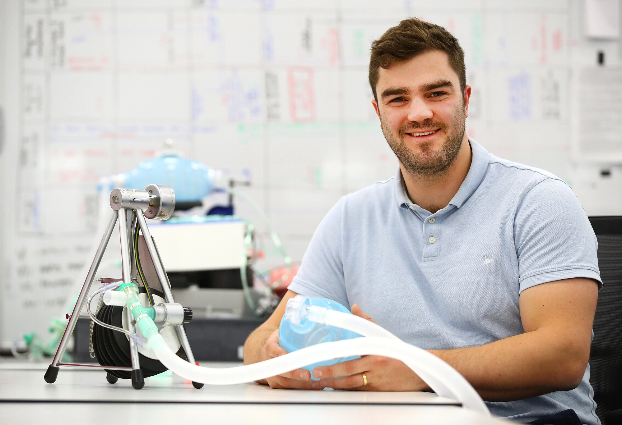 Man in a light blue shirt smiling, seated at a desk with a medical ventilation device, whiteboard in the background.